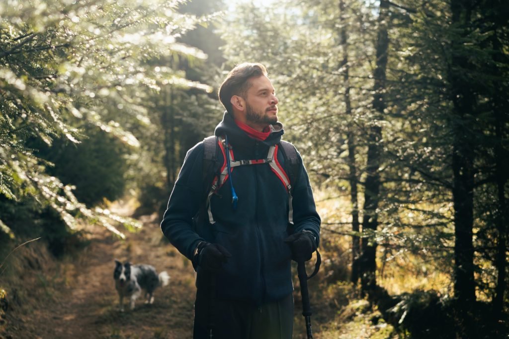 Hiker with backpack walking in a sunlit forest with a dog in the background.